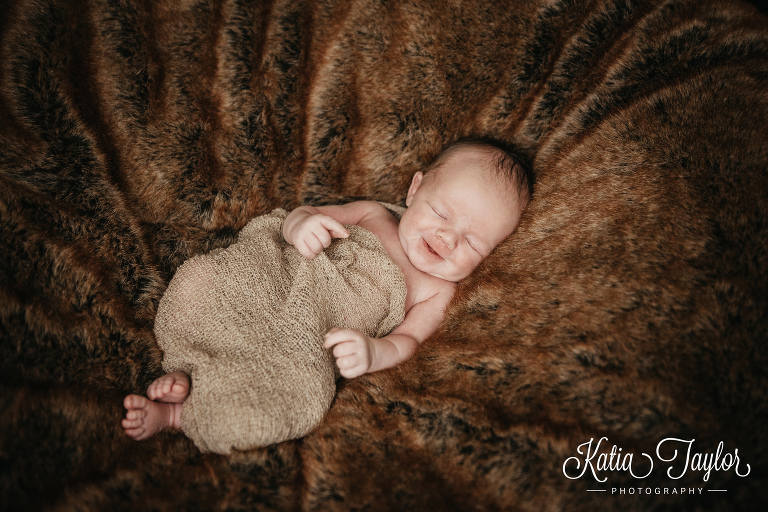 Newborn baby boy smiling in beige wrap on fur blanket. Toronto newborn baby photographer.