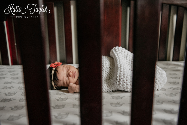 Newborn baby girl asleep in her crib in a white wrap with orange flower headband. Toronto newborn portraits.