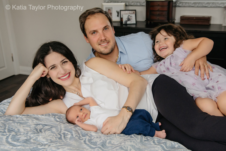 Family of four with a newborn baby boy and a toddler girl laughing while playing on the bed
