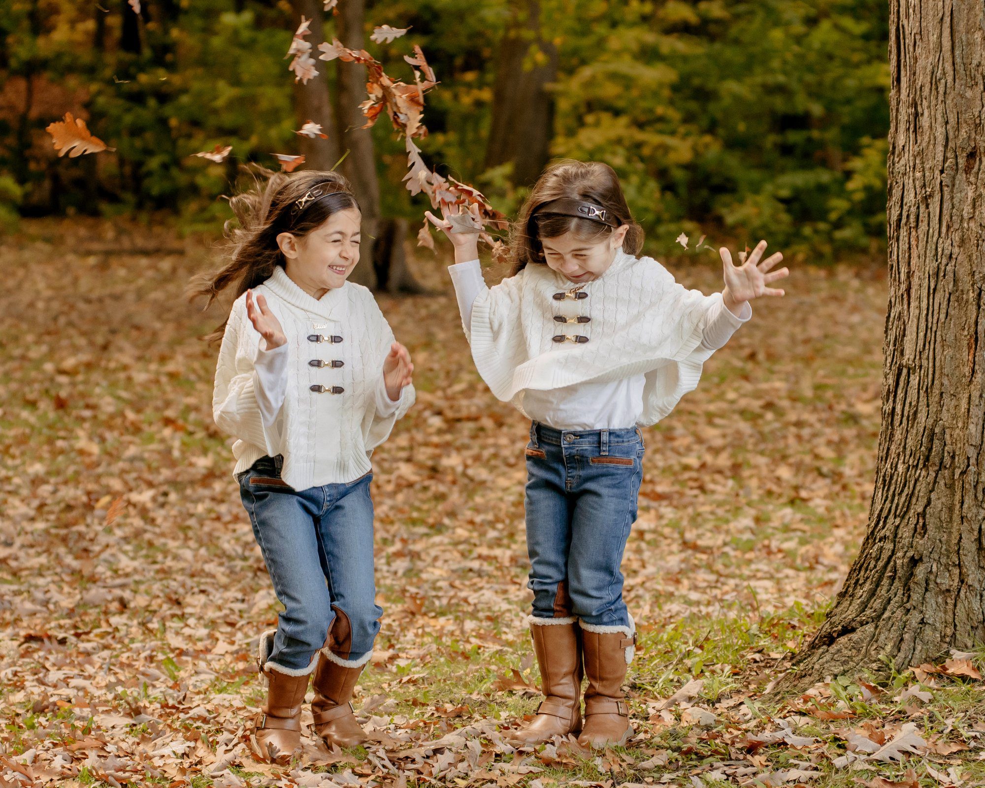 Two young sisters in white ponchos and blue jeans throwing leaves in the fall in a park in Toronto
