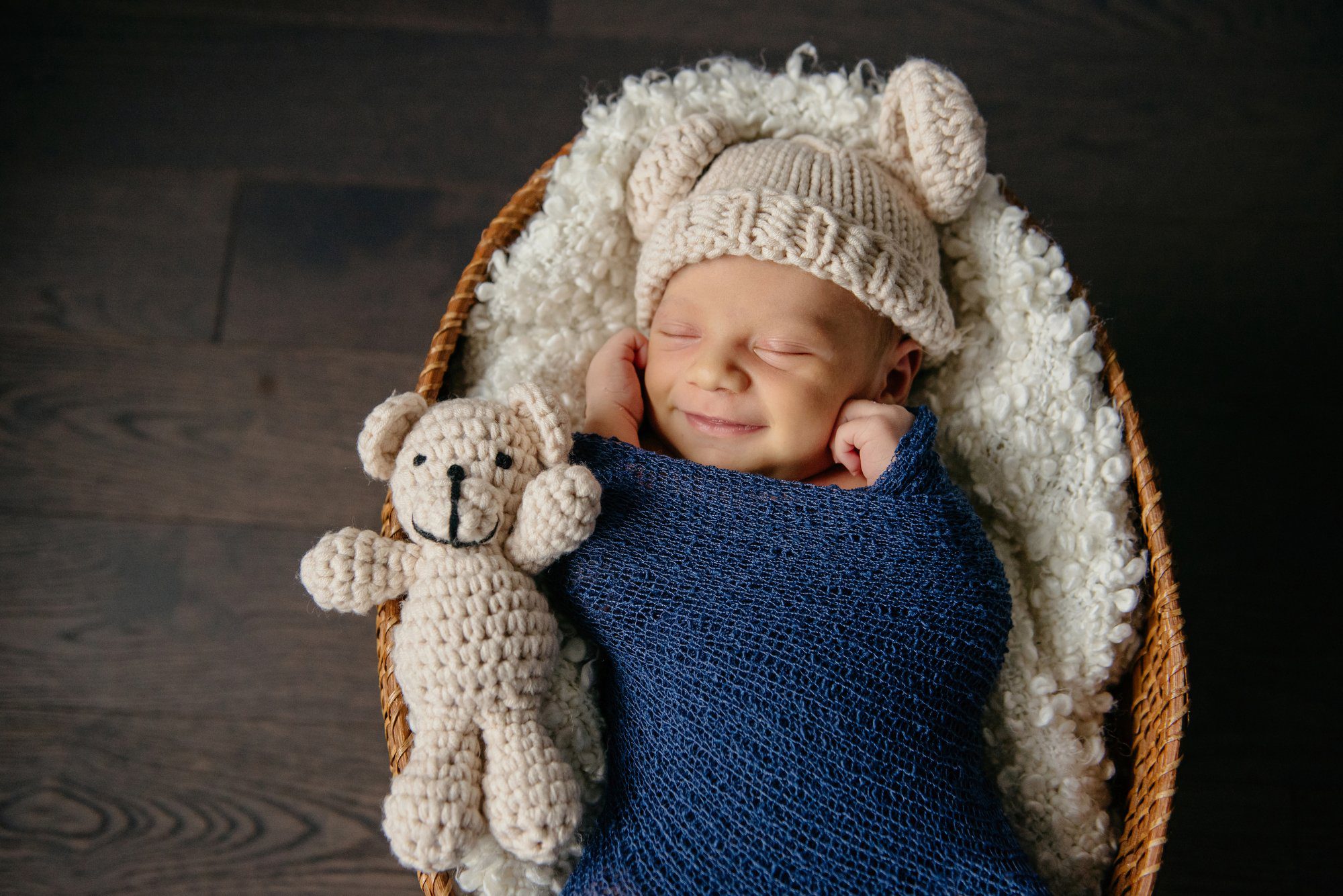 An adorable newborn baby boy smiles in his sleep wearing a crocheted bear eared hat with teddy bear